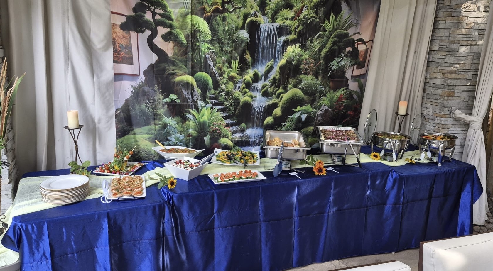 Indoor buffet catering spread with chafing dishes and appetizers against a nature backdrop