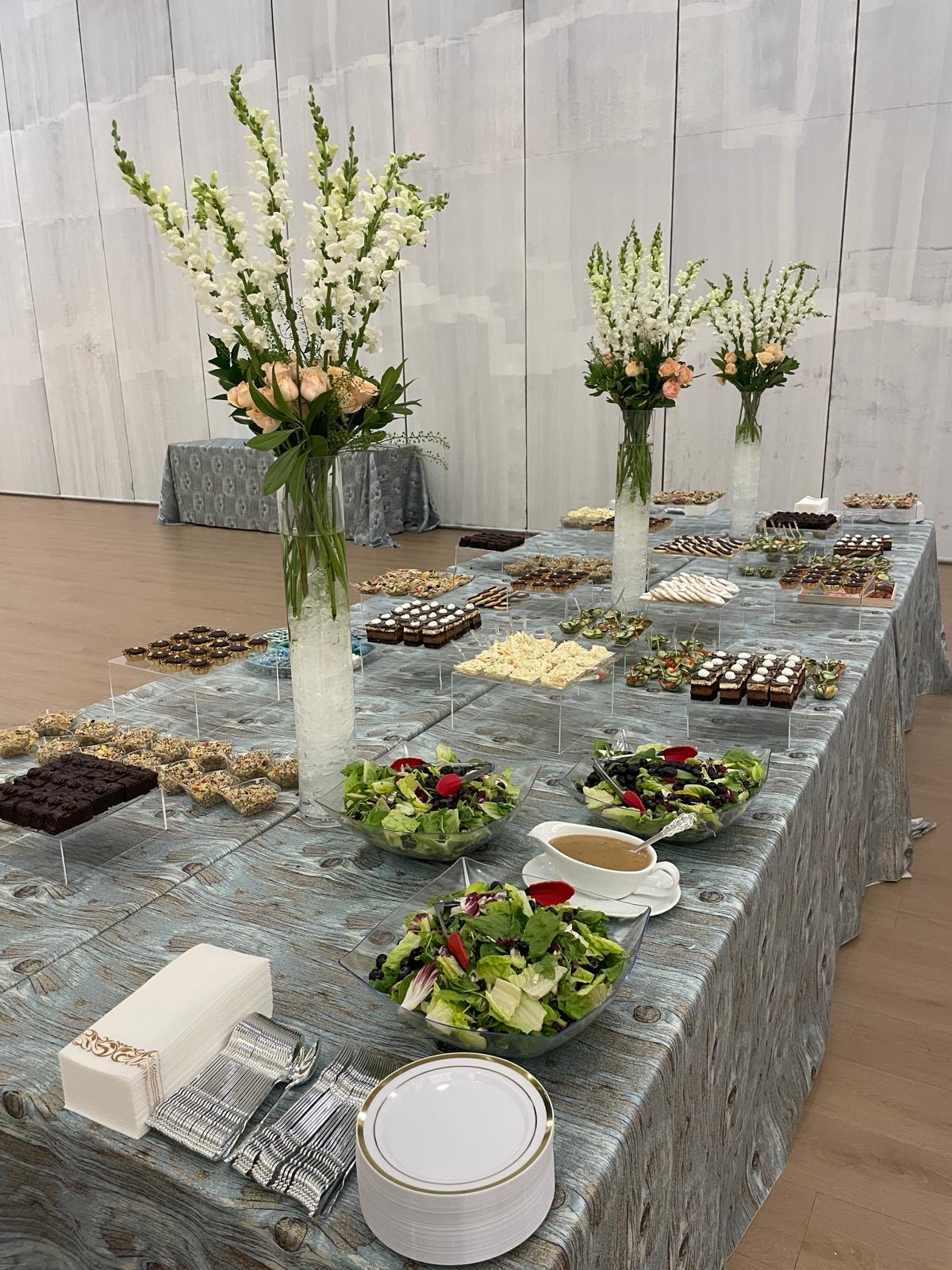 Elegant catering table with tall floral arrangements, dessert platters, and fresh salads